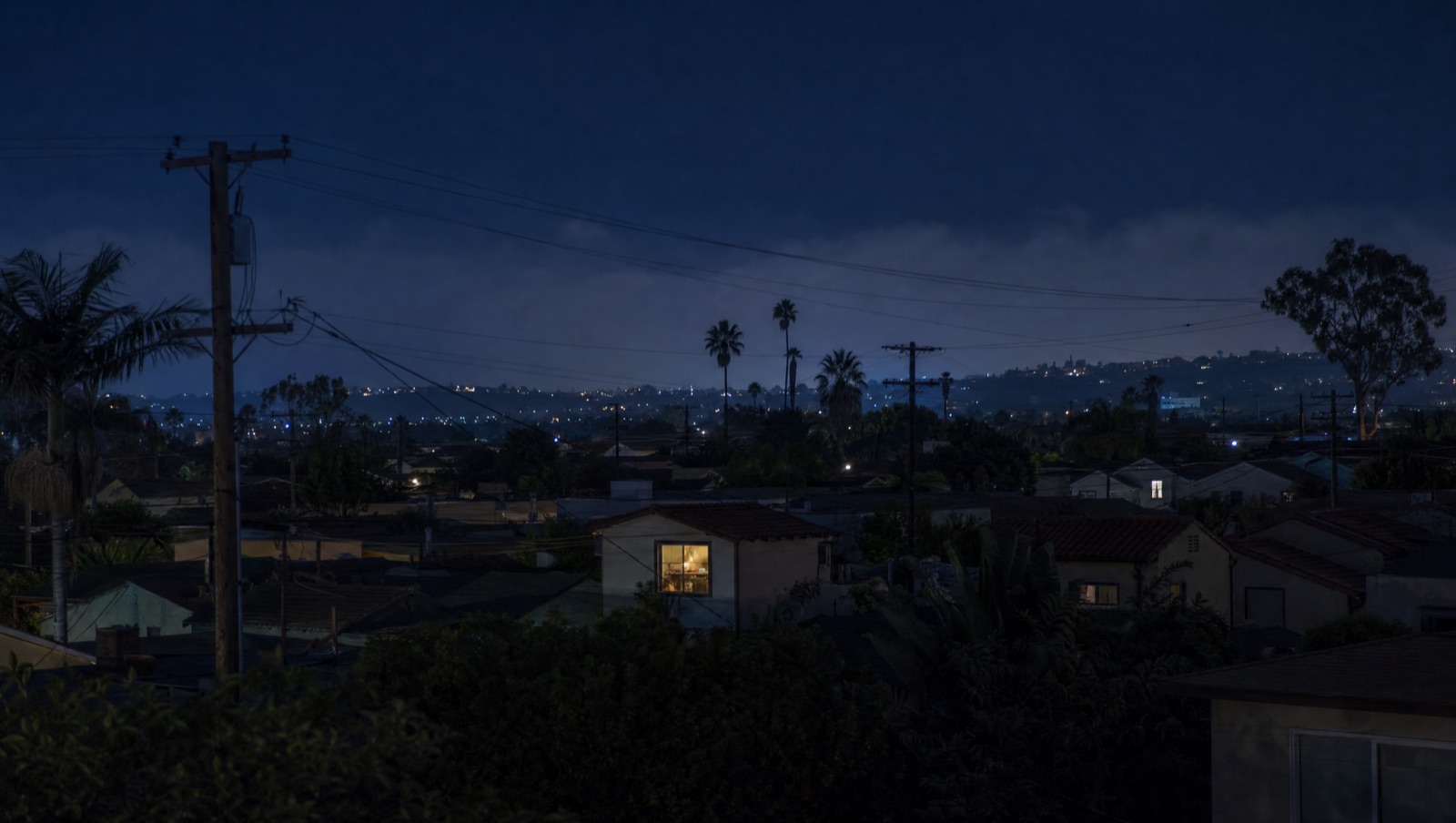 San Diego rooftops at night under a deep blue sky, one kitchen window glowing warm